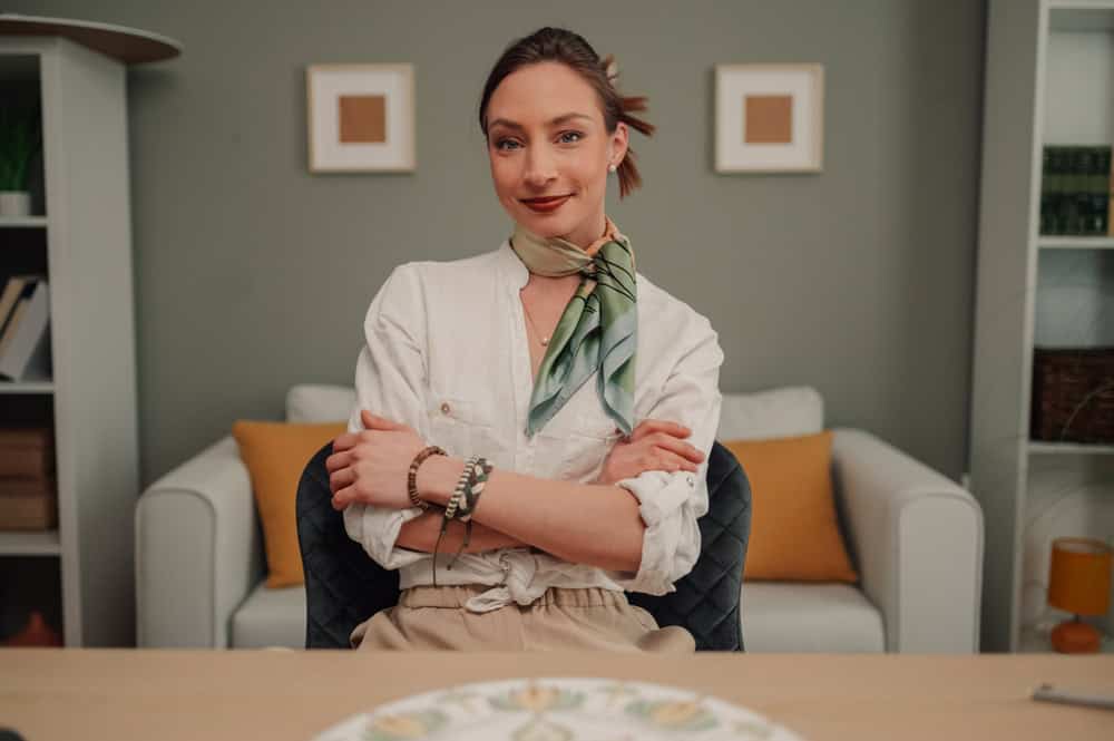 Businesswoman wearing a silk scarf and bracelets sits with folded arms at her desk in a stylish home office, exuding confidence and professionalism