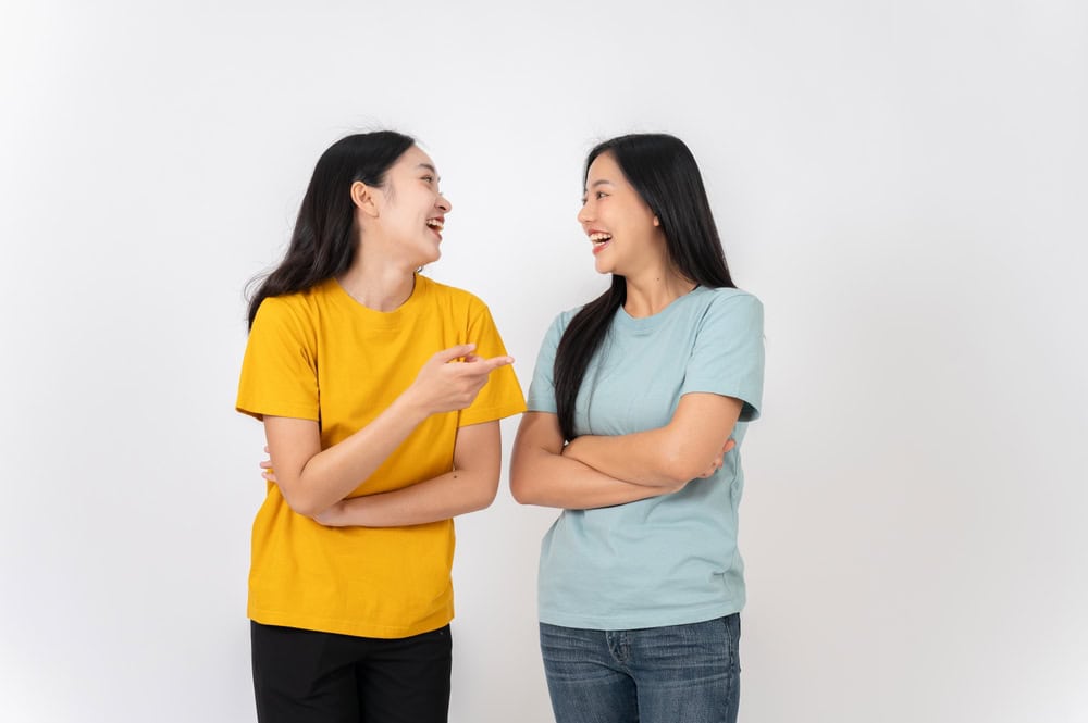 Two cheerful, lively Asian female friends in casual clothes standing against a white studio background, enjoying talking and laughing together. best friend, friendship, relationship