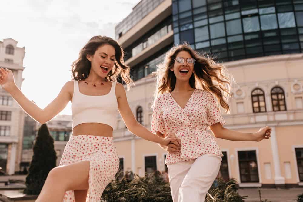 Funny fair-skinned girls with open mouths actively move in fresh air. Brunette in white top and skirt with slit. Blonde is dressed in blouse and white pants, in transparent glasses.