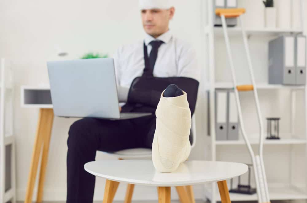 Businessman with a broken leg, wrapped in a bandage, working on laptop in an office. Injury and trauma from the accident, he remains dedicated to work, showing resilience and determination.