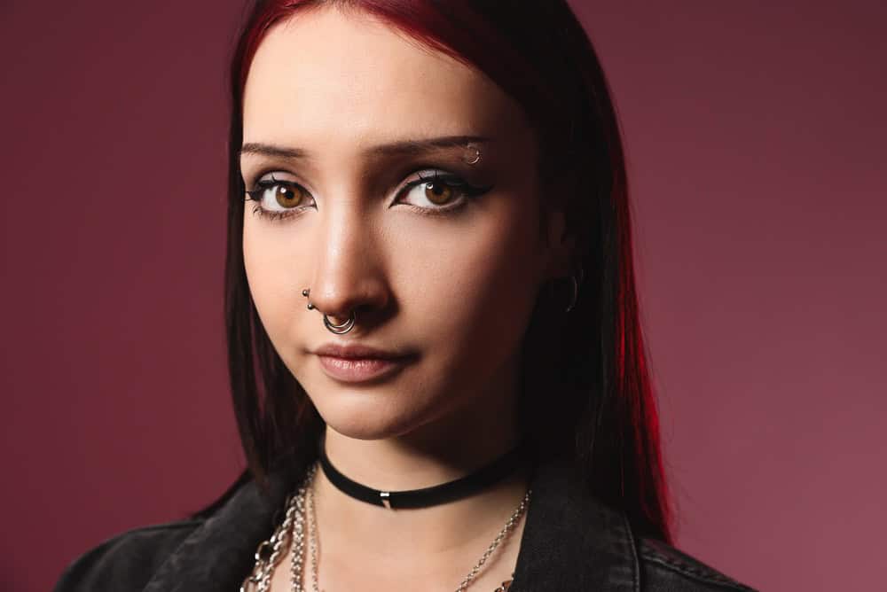 Portrait of beautiful young woman with different face piercings and choker on pink background