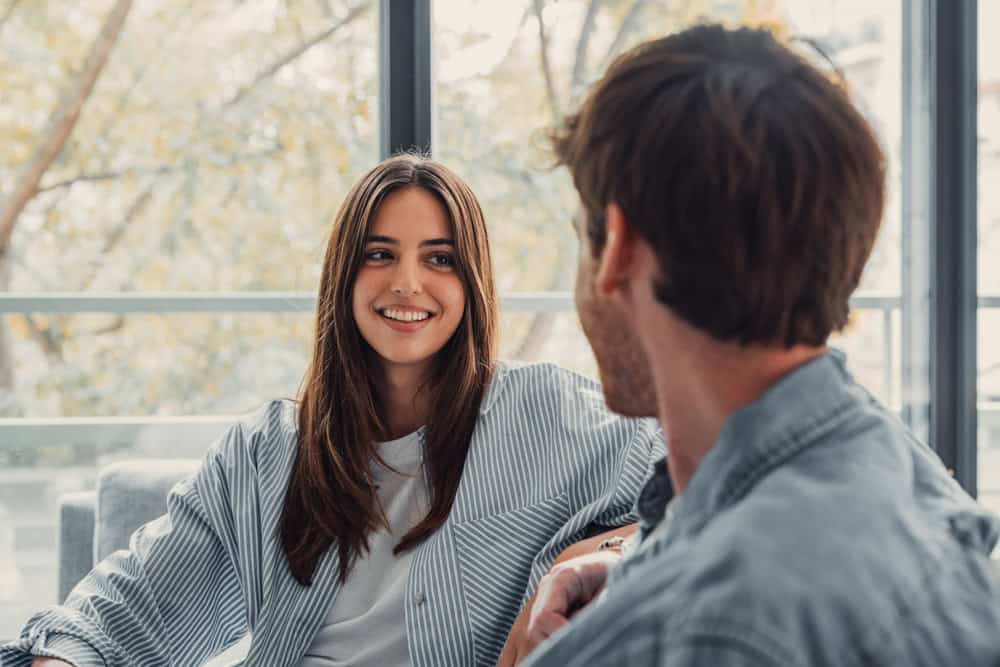 Young happy couple having fun talking laughing relaxing at home on couch, boyfriend embracing girlfriend telling funny joke sitting on sofa.