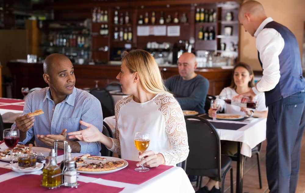 Positive interracial married couple sitting in cozy restaurant and eating pizza with wine