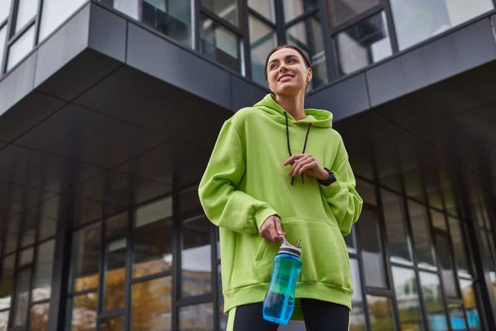 happy sportswoman in oversized hoodie and leggings standing with bottle with water outdoors