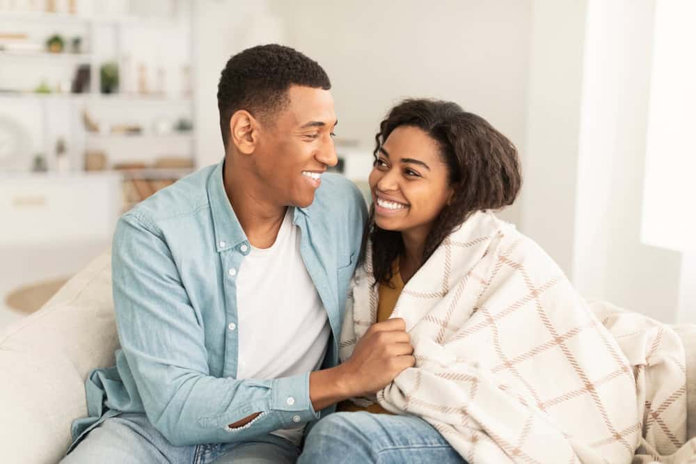 Happy young african american male covers with blanket and hugs his wife, sit on sofa, enjoy spare ti