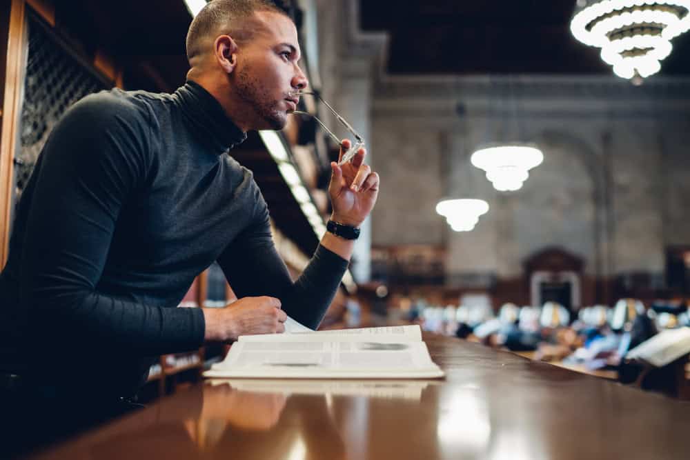 Smart male pondering on information from poetry sitting in book archive at late evening, thoughtful serious young academic professor in library thinking on readed material of classics literature