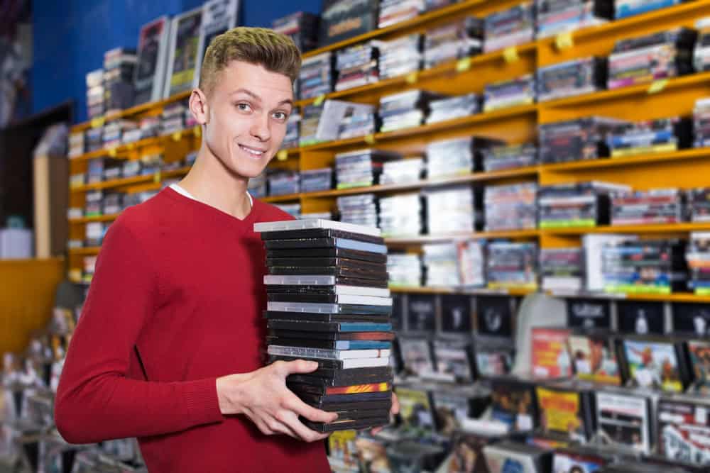 Positive young guy holding pile of boxes with DVDs bought in shop