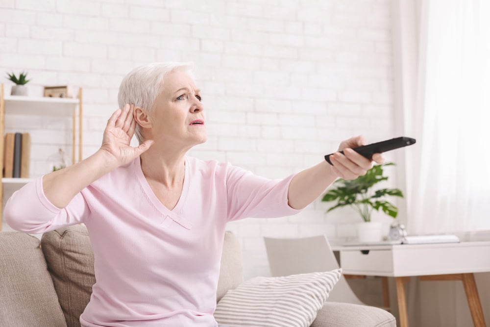Senior woman cupping her hand behind ear to hear better to the television, making sound louder, empty space