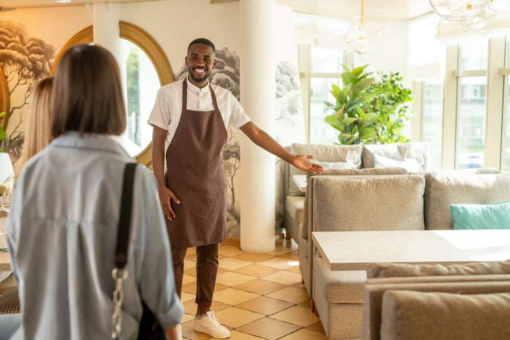 Young cheerful waiter in workwear inviting female guest to take place by one of tables surrounded by soft comfortable couches in restaurant