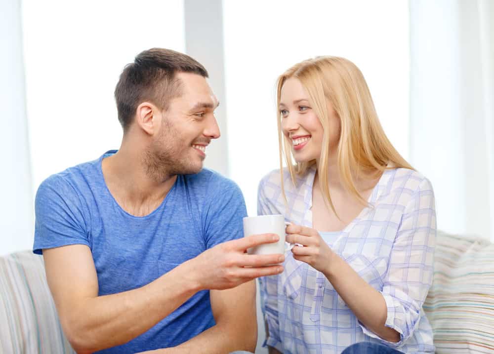 Love, family, healthy food and happiness concept - smiling man giving cup of tea or coffee to wife or girlfriend at home