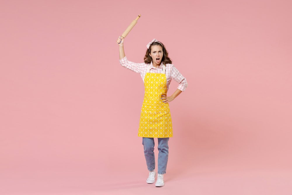 Full length portrait of angry young brunette woman housewife 20s in yellow apron hold rolling pin swearing while doing housework isolated on pastel pink colour background studio. Housekeeping concept