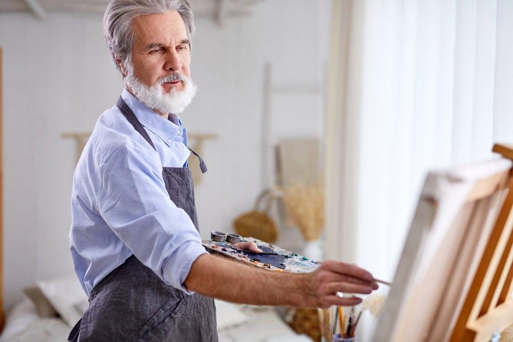 artist holding creating masterpiece using paintbrush, stand near canvas, gray haired male in apron enjoy drawing