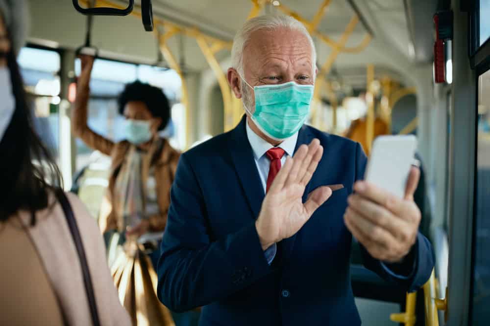 Happy senior businessman waving during video call while commuting by bus during coronavirus pandemic.