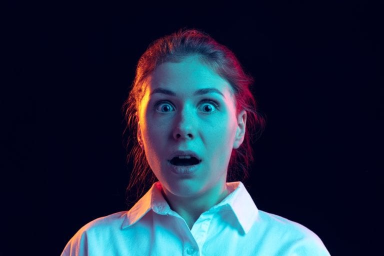 Shocked young girl in white shirt looking at camera with open mouth isolated on dark background in blue neon light, filter. Concept of emotions, facial expression, youth, aspiration, sales.