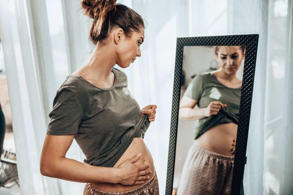Unhappy woman standing in front of a mirror and holding her stomach while suffering from cramps.