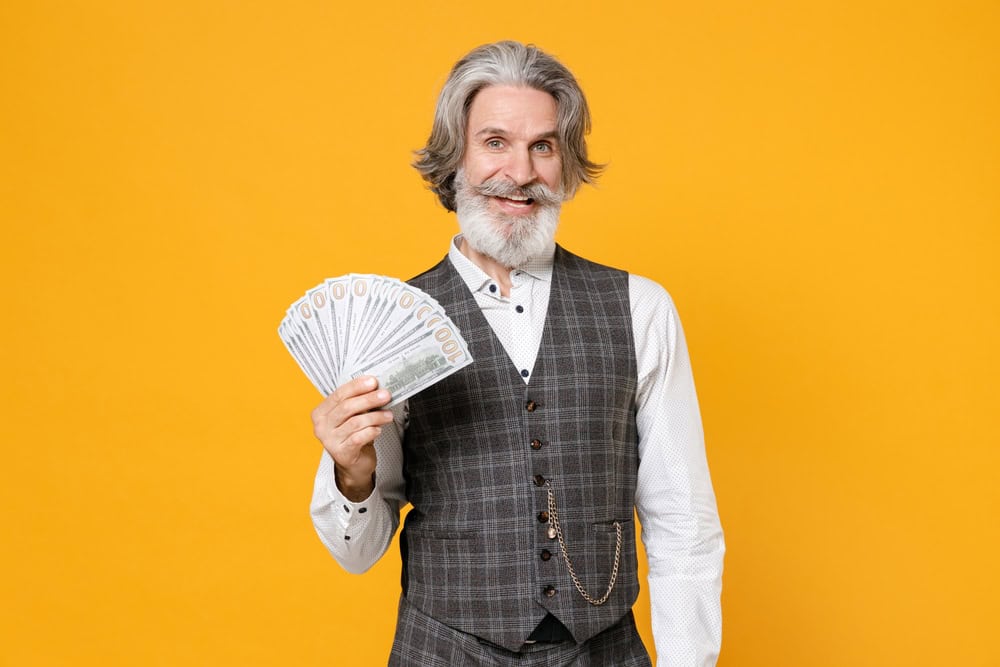 Smiling elderly gray-haired business man in checkered suit waistcoat shirt hold fan of cash money in dollar banknotes isolated on yellow background studio portrait. Achievement career wealth concept