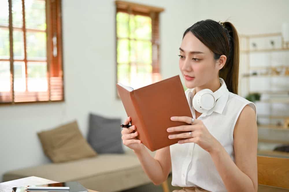 Beautiful and focused millennial Asian woman checking her schedule on her planner book, reading a book in her minimal living room.
