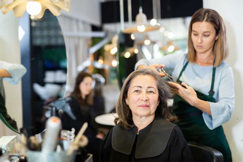 Elderly silver haired woman getting haircut by professional young female hairdresser in modern hair salon.
