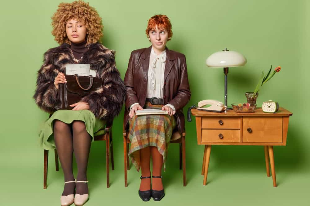 Two women pose on chairs wait for job interview wear fashionable retro clothes pose with papers pose in room with old furniture isolated over green background. Elegant female models being cabinet