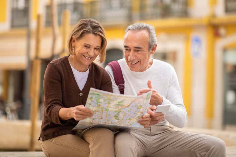 Ageless Wanderlust. Smiling Senior Couple Holding Touristic Map Planning Their Summer Vacation And Choosing Travel Destination Sitting Outdoor. Travelers Spouses Using Paper Map For Navigation