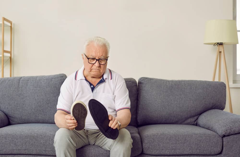 Puzzled funny gray-haired elderly man holds at his shoes in his hands, sitting on green sofa at home. Cute confused grandpa in white polo shirt chooses shoes for significant festive event.