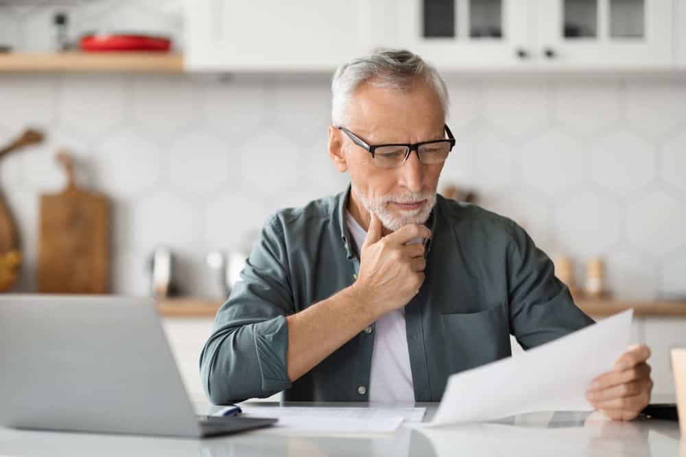 Paperwork Concept. Portrait of senior man working at home office, holding and reading papers or financial document, sitting at desk in kitchen, using pc laptop computer for remote work, closeup shot