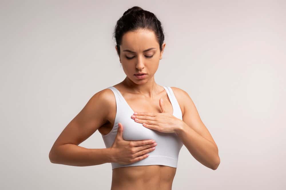 Breast screening. Young brunette woman examining her breasts, wearing white top, cancer prevention concept, isolated on grey background, closeup, copy space