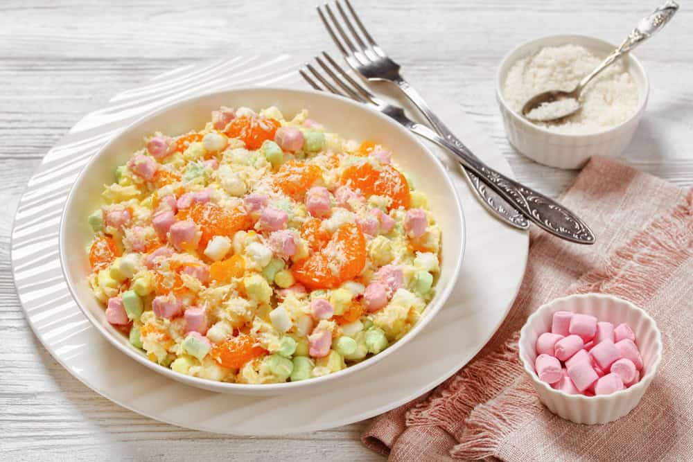 Ambrosia Salad with pineapple, mandarin oranges, yogurt, mini marshmallows, coconut and whipped cream in white bowl on white wooden table, american recipe, horizontal view