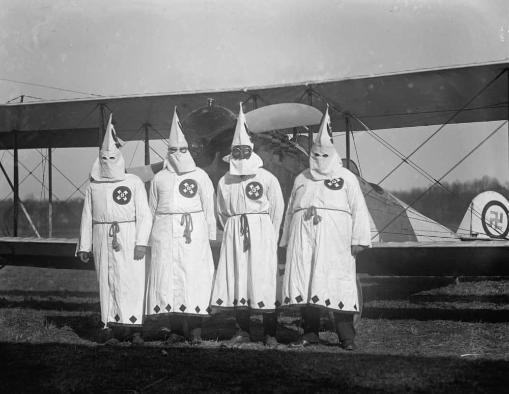 Four hooded Ku Klux Klan members pose in from of their airplane which bears a swastika emblem. 1922 photo.