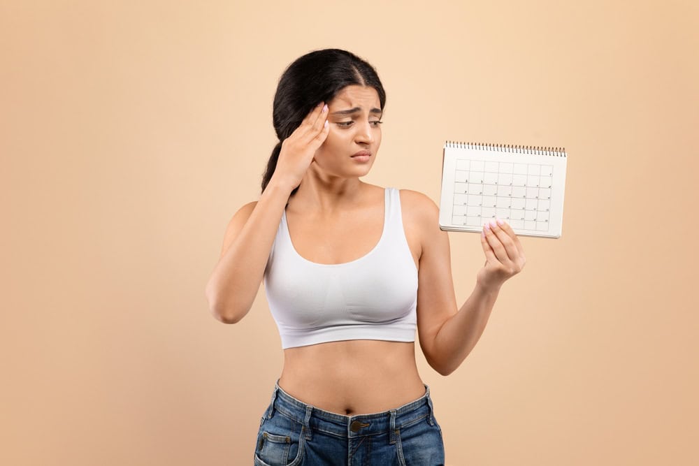 Concerned young indian woman holding blank menstrual cycle calendar, upset eastern female tracking her monthly period, indicating stress about irregularities, standing on beige background