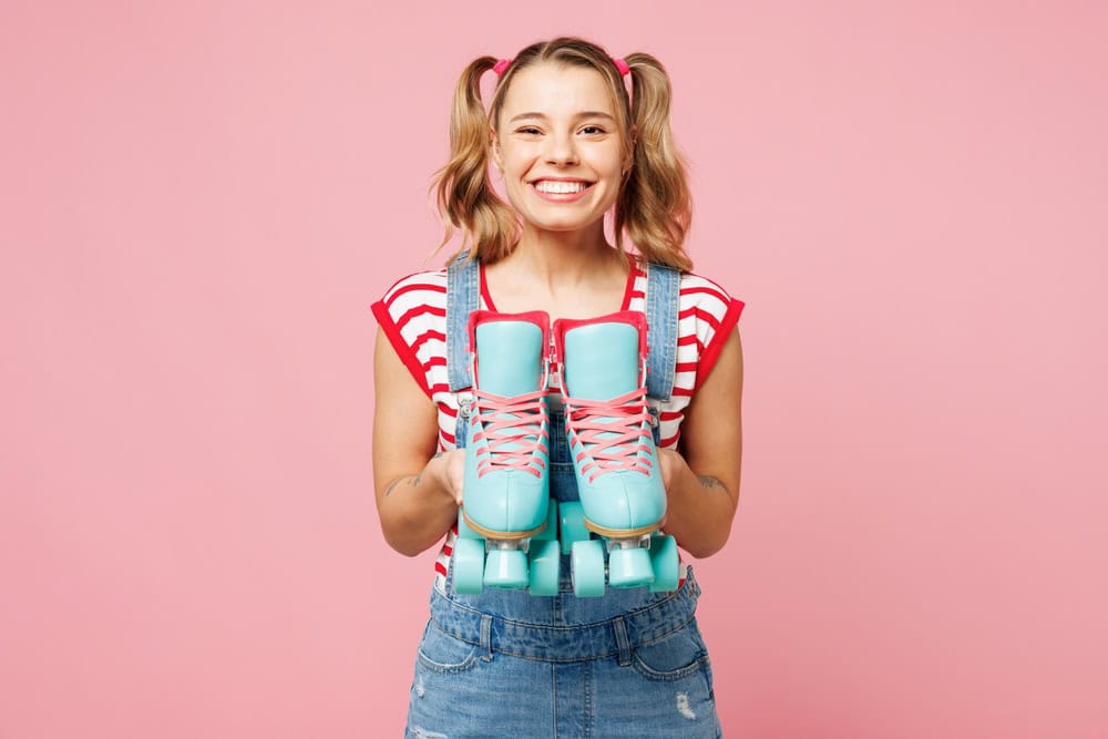 Young smiling happy fun woman wear red t-shirt denim overalls casual clothes hold in hand give blue rollers look camera isolated on plain pastel pink background. Summer sport lifestyle leisure concept