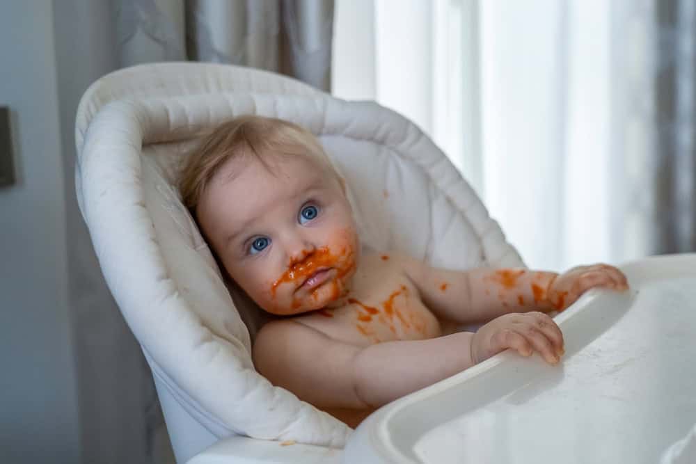 Adorable baby girl sits in a high chair with food smeared all over their face and body