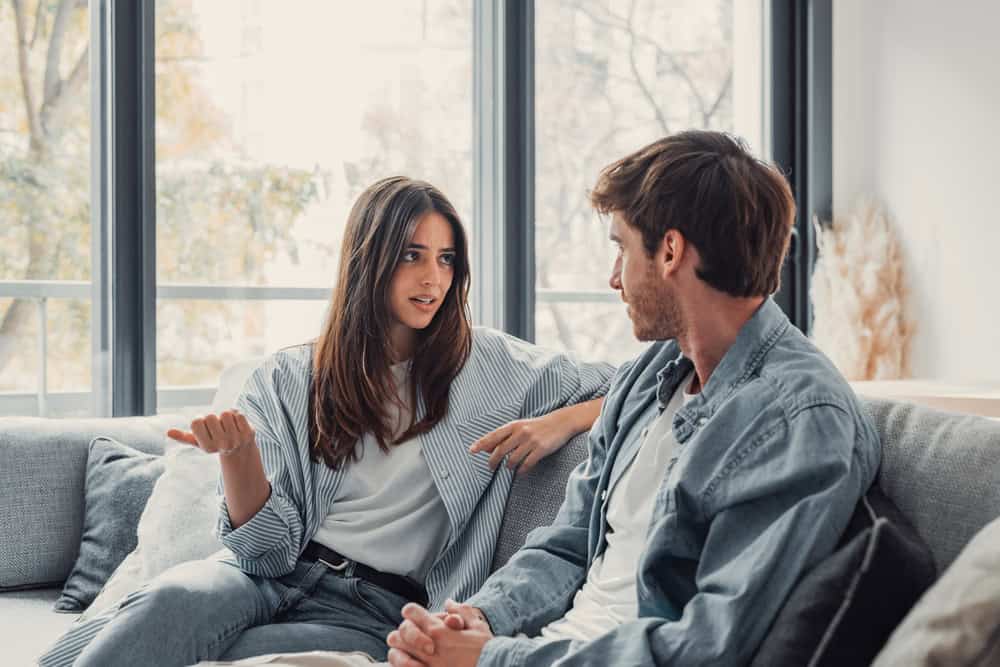 Young happy couple having fun talking laughing relaxing at home on couch, boyfriend embracing girlfriend telling funny joke sitting on sofa, humor in relationships, enjoying weekend together
