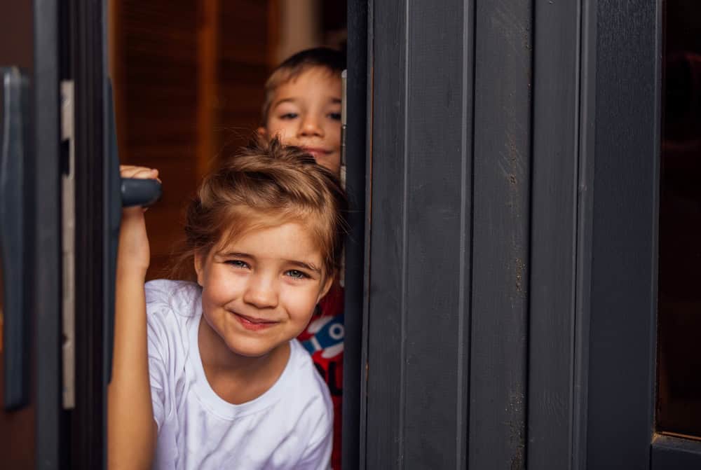Close-up of cute children peeking out of the doorway. Little brother and sister open the front door. Cute kids meet their parents or guests. A boy and a girl have fun together at home.
