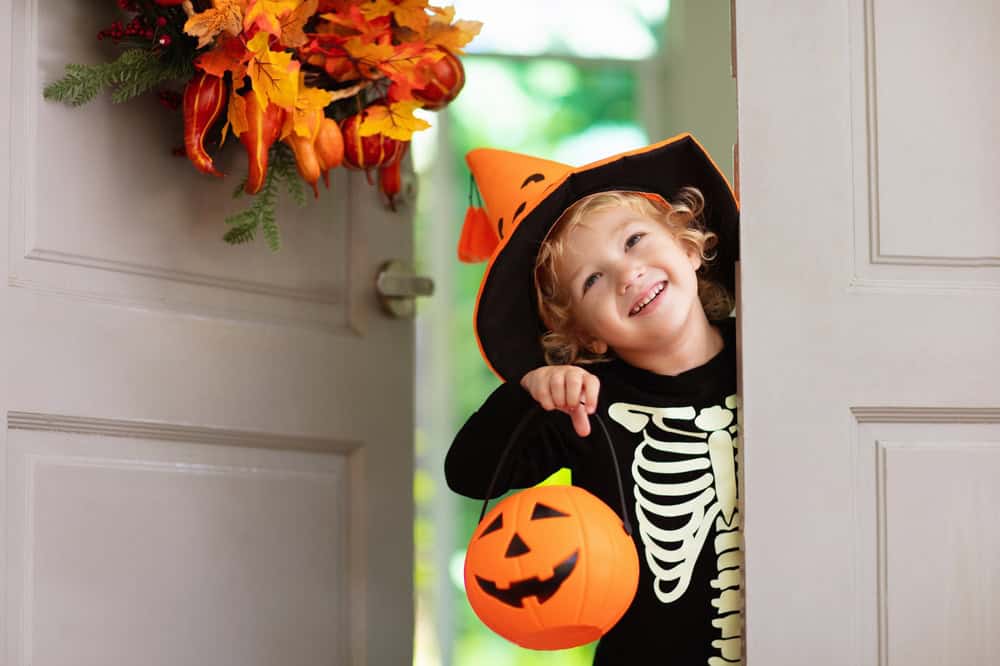 Kids trick or treat on Halloween night. Child at decorated house door with autumn leaf wreath and pumpkin lantern. Little boy in witch and skeleton costume and hat with candy bucket. Fall decoration.
