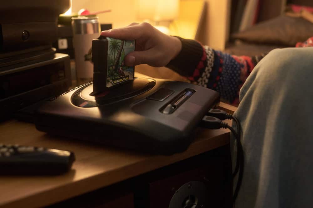 Person inserting video game cartridge into classic console while sitting in cozy room with warm lighting. Remote control and other retro devices can be seen on table