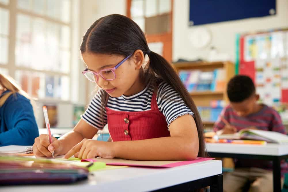 Concentrated cute little primary schoolgirl writing in book with classmates in background at classroom. Little smart pupil writing at desk in classroom at the elementary school.