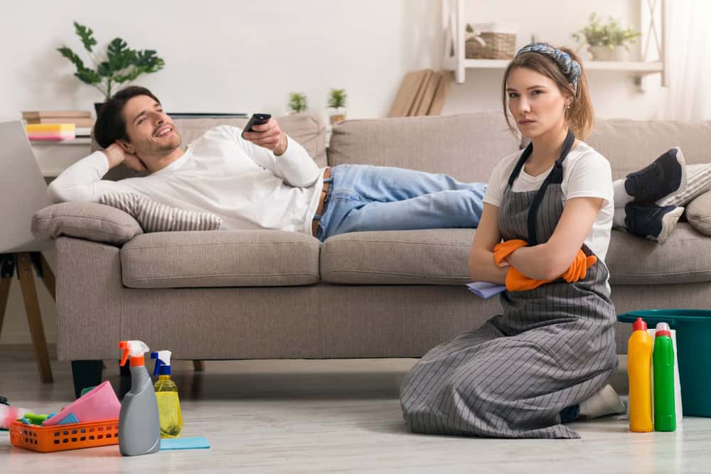 Household Chores Problem. Offended Woman Tired After Cleaning Sitting Next To Lazy Husband Relaxing On Couch
