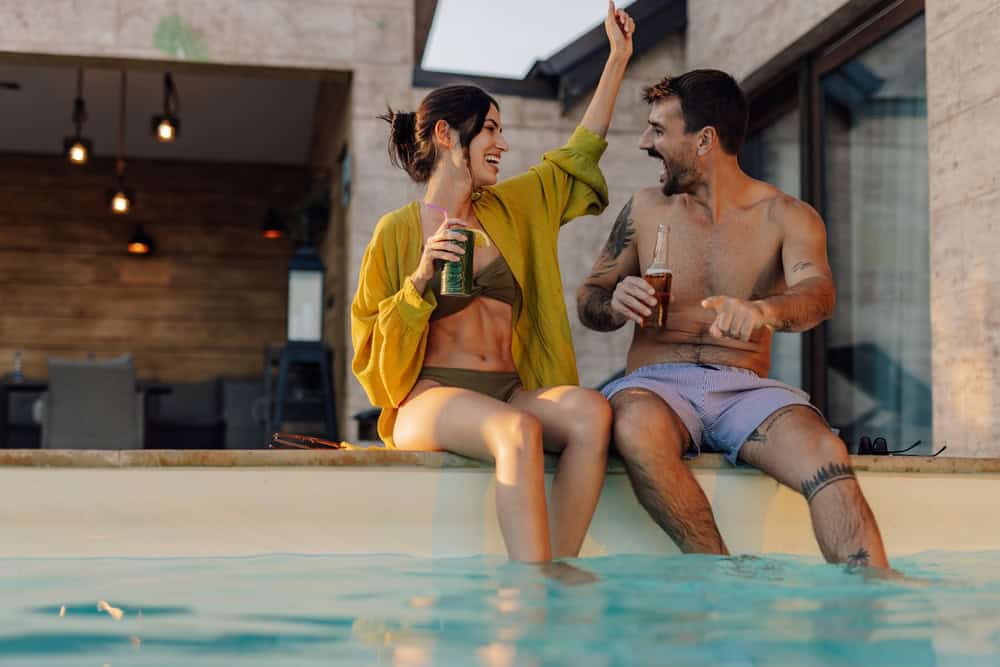 Happy couple sitting by swimming pool, holding drinks, woman raising one arm and laughing, man pointing and laughing, during summer vacation