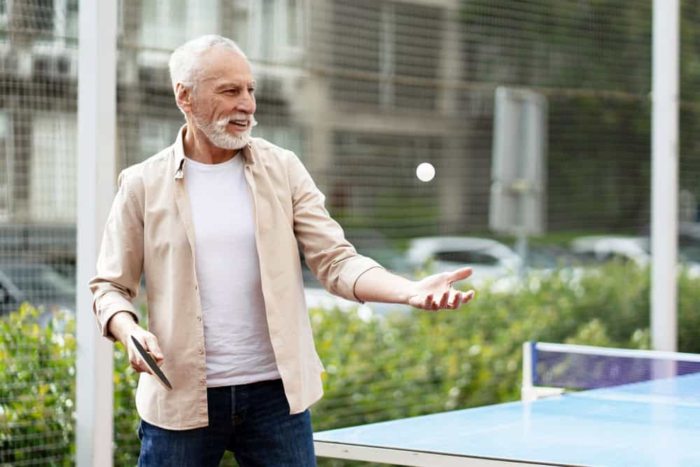 Elderly man enjoying a game of ping pong in the park, skillfully throwing a ball up to serve while smiling and staying active outdoors