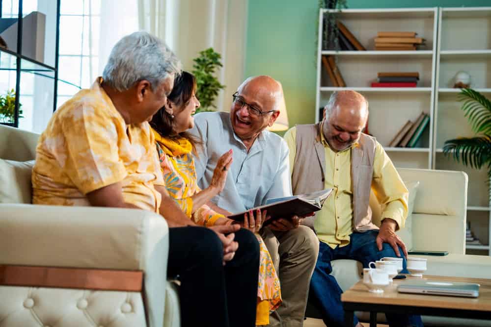 Seniors looking at photo album while seated on cozy sofa indoors, Indian Asian elderly friends flipping through cherished pictures during emotional joyful reunion at modern home living room together