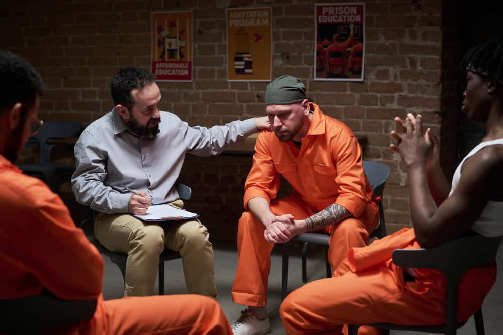Caucasian man in orange prison uniform sitting with head down, while young adult Caucasian man with clipboard comforting him during group therapy session with inmates