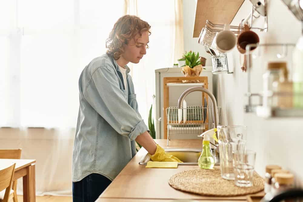 A young man with curly hair washes dishes in a warm kitchen filled with natural light.