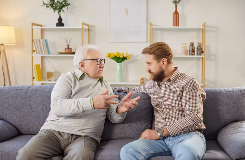 Young son and elderly father having conversation. White haired old man in his 60s or 70s sitting on sofa at home together with adult son, talking about life and sharing advice. Senior parents concept