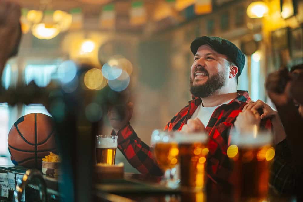 Happy bearded man cheering with beer in hand while watching basketball at a lively pub with friends, smiling and celebrating victory