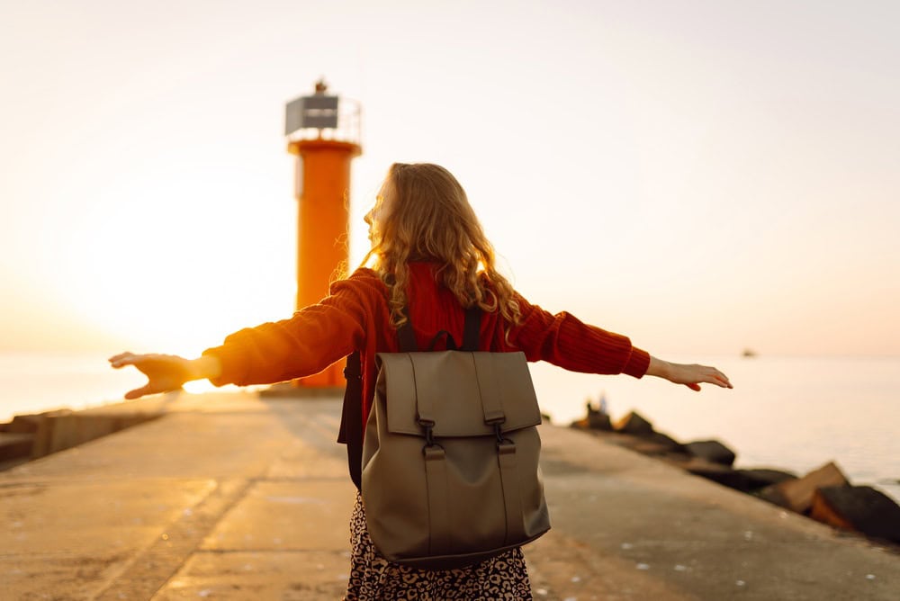 A happy woman enjoys a walk along a pier near a lighthouse at sunset. A backpacker feels free at sunset. Concept of nature, freedom, and adventure.