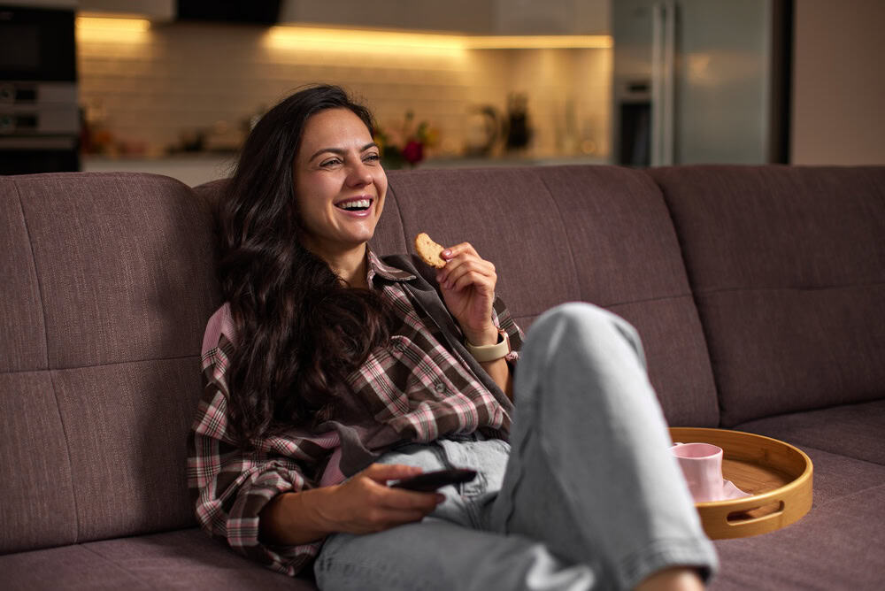 Woman feeling happy, laughing and relaxing on a sofa at home, eating a cookie and watching tv in the evening
