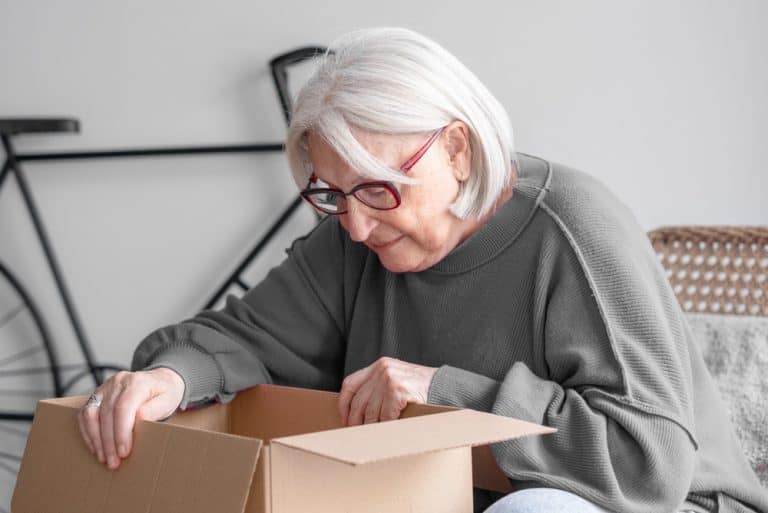 Senior woman opening delivery box at home, exploring new purchases, experiencing online shopping convenience, receiving package with curiosity and surprise