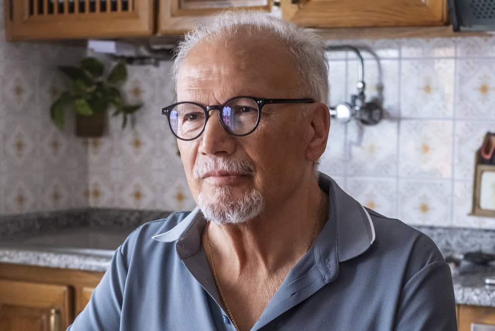 Senior man with white hair and glasses thinking, looking into the distance, reflecting on future plans or past memories while standing in a home kitchen setting