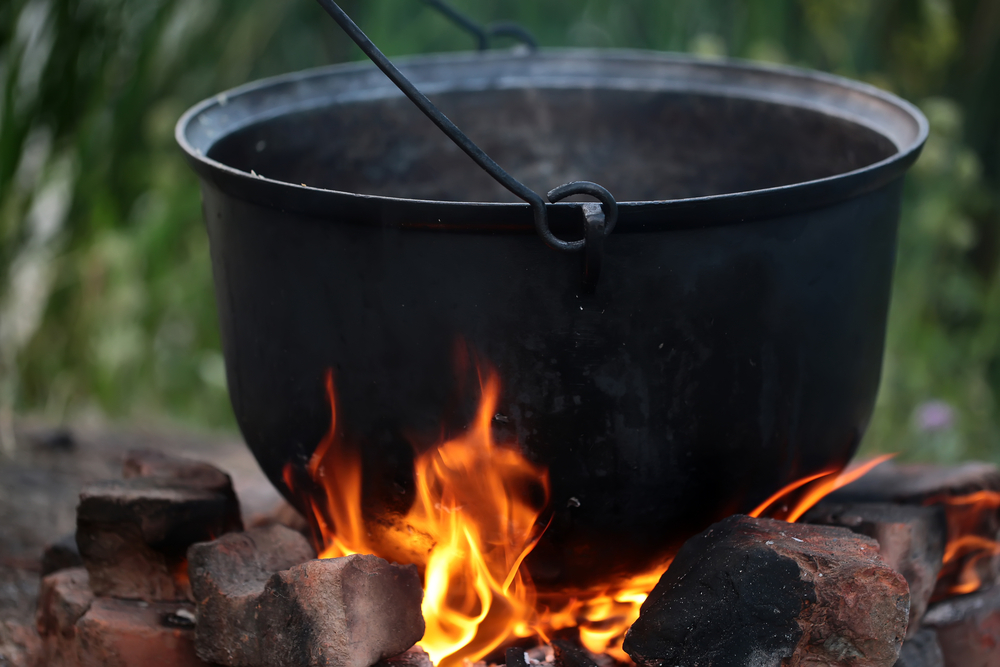 Closeup cooking in cauldron licked by flames on open fire fireplace made of bricks stones in field conditions over blurred nature background, horizontal picture